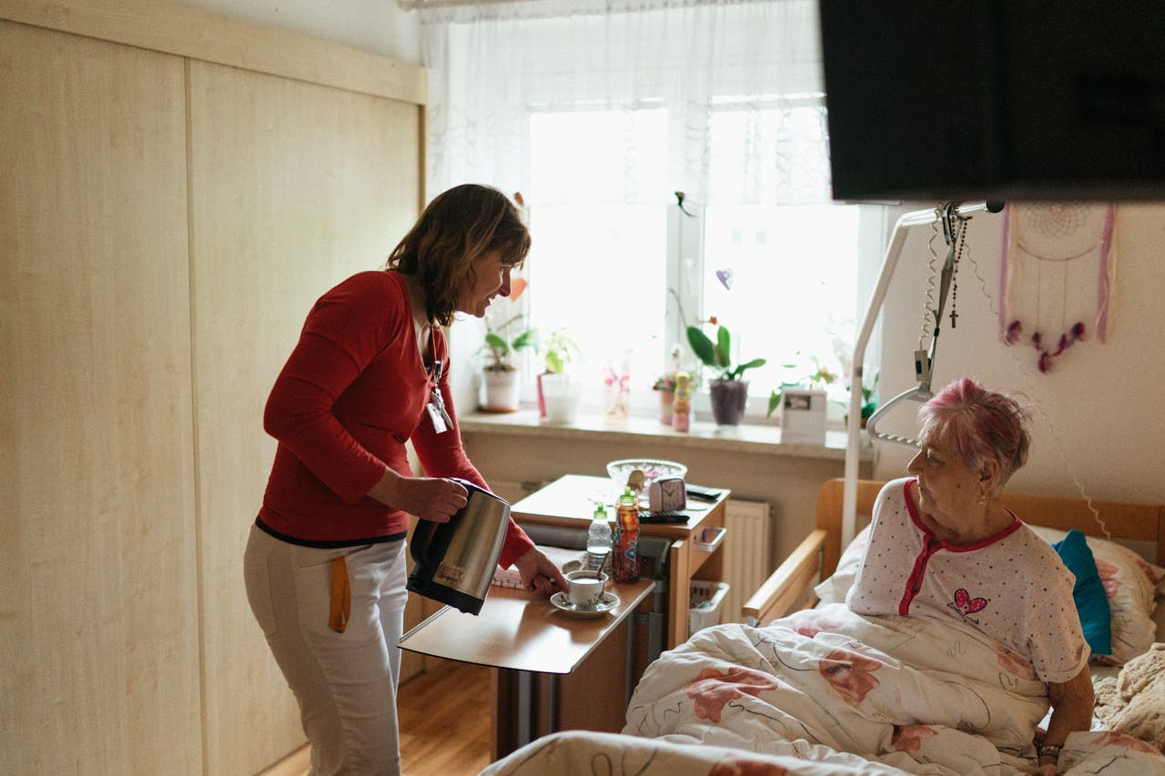 our-journey-02 A caregiver serves tea to a senior woman in a cozy bedroom setting, fostering comfort and care.