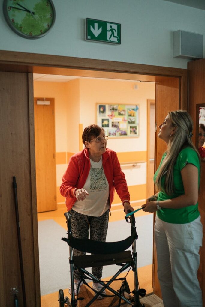 A caregiver assists a senior woman with a walker in a retirement home corridor.