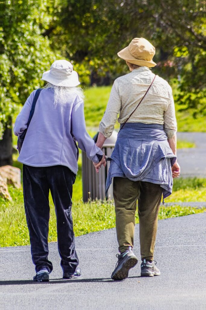 Two elderly women stroll hand in hand on a sunny day in the park, wearing hats.