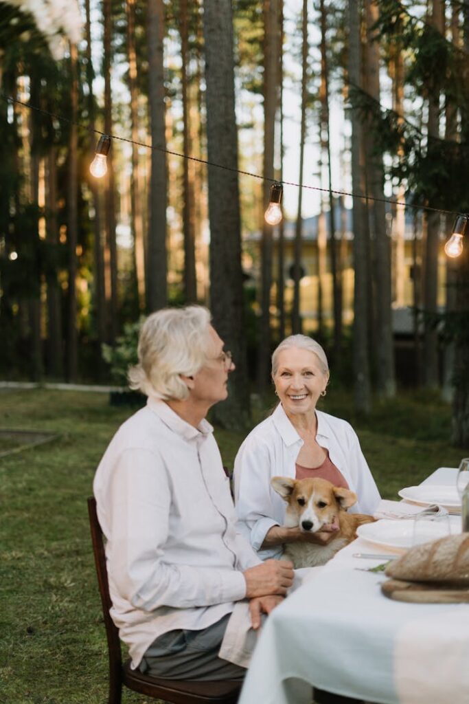 Elderly couple dining outdoors with a corgi, embracing nature and happiness.