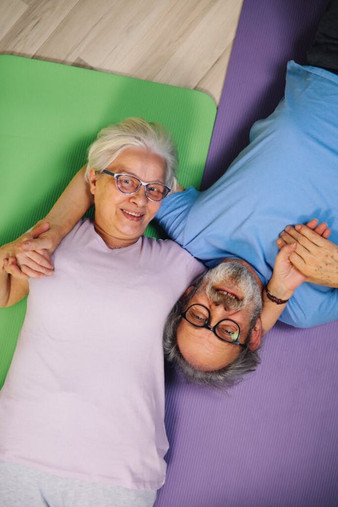 Elderly couple smiling and holding hands while lying on yoga mats indoors.